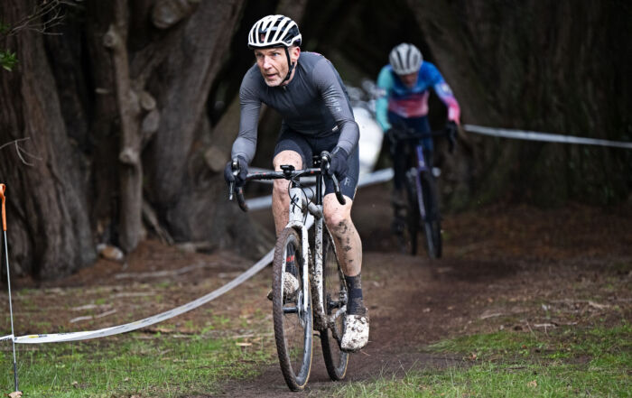 Two cyclists ride on a muddy forest trail during a race, with one rider in gray leading and another in blue and pink behind, both benefiting from INSCYD testing for cyclists beneath arching trees.