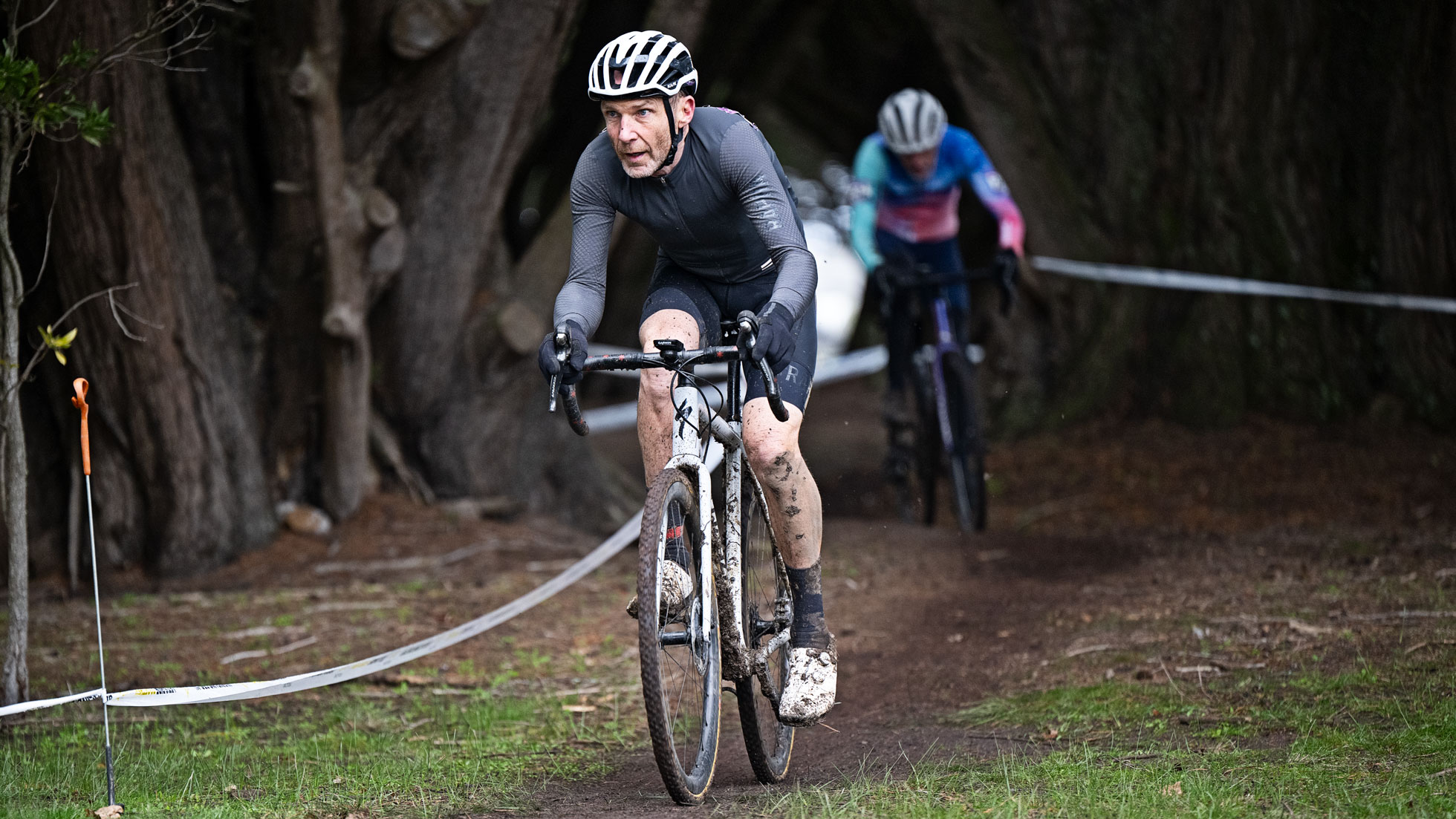 Two cyclists ride on a muddy forest trail during a race, with one rider in gray leading and another in blue and pink behind, both benefiting from INSCYD testing for cyclists beneath arching trees.