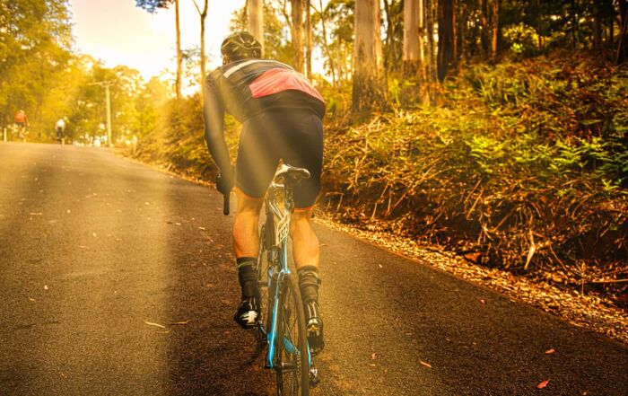 A cyclist rides uphill on a sunlit road through a forest, training for the Peaks Challenge Falls Creek, with beams of sunlight streaming through the trees and casting a warm, golden glow. Another cyclist is visible ahead in the distance.